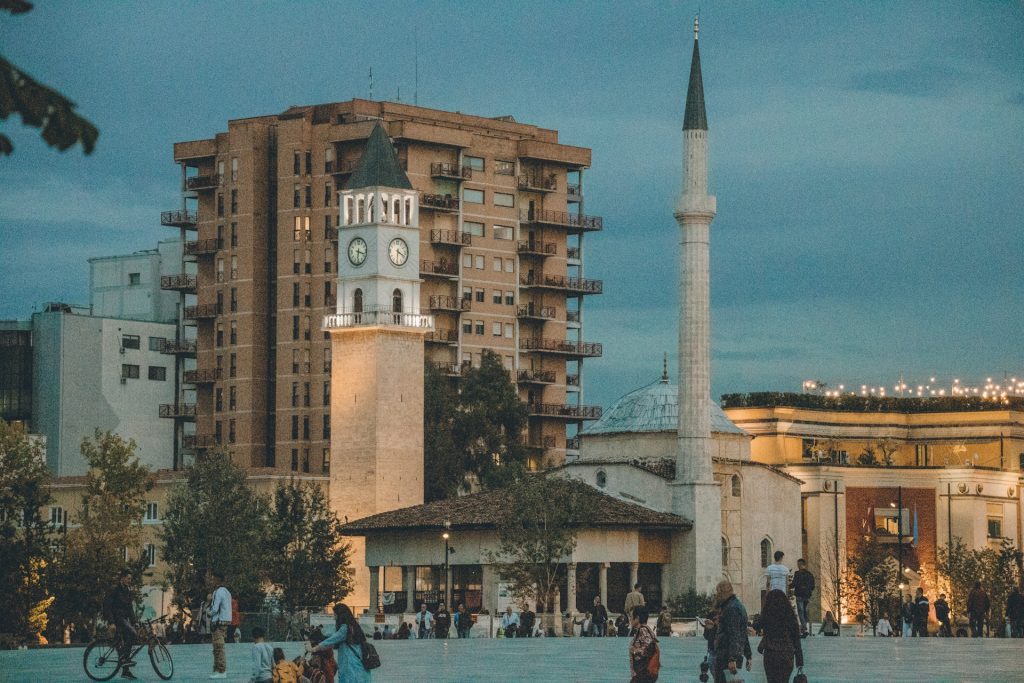 a group of people walking around a plaza with a clock tower in the background